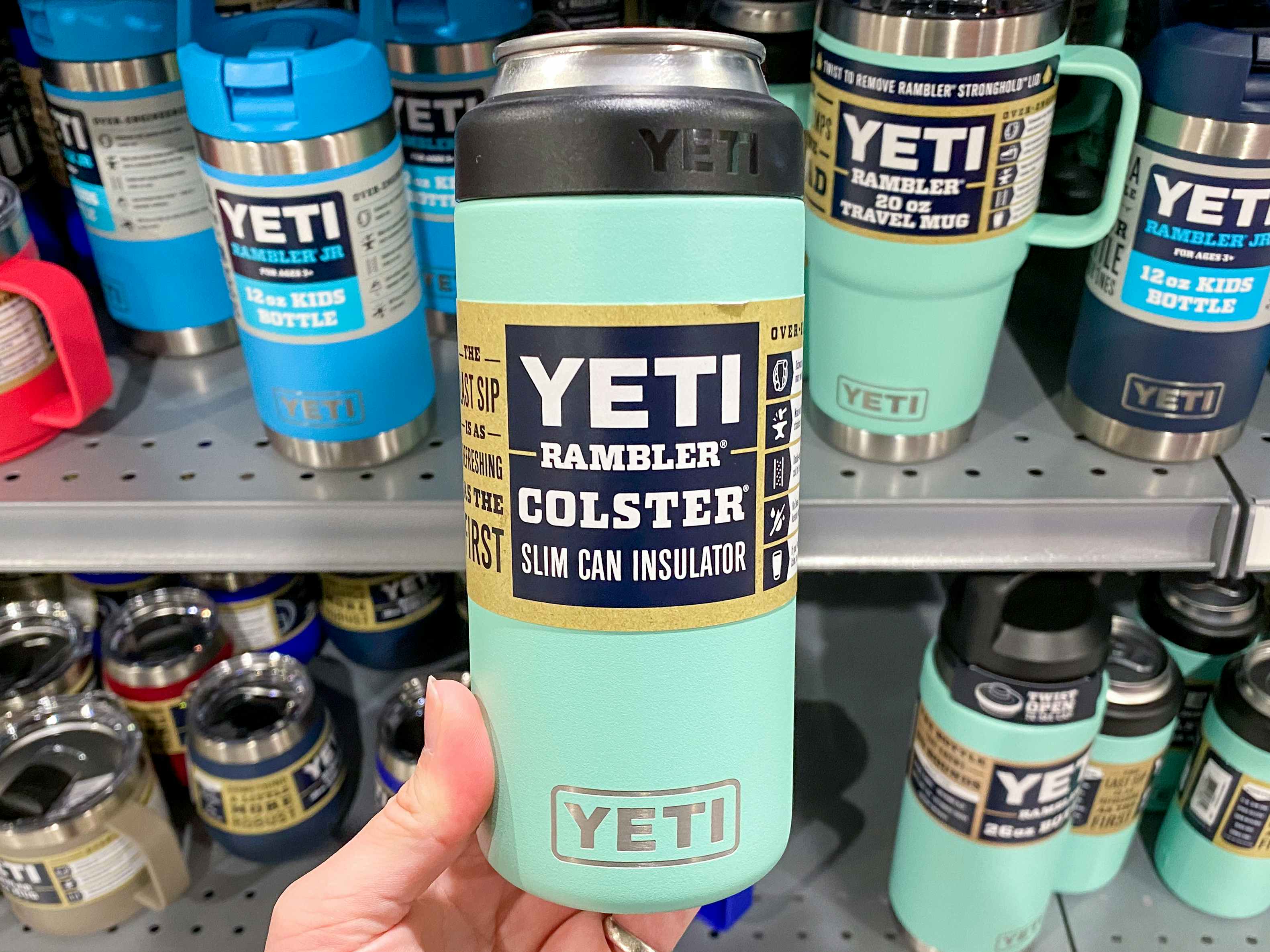A person's hand holding up a teal Yeti Rambler Colster Slim Can Insulator in front of a shelf of more Yeti products at Dick's Sporting ...