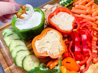 a hand dipping a pretzel into dip from a bell pepper filled with dips on a veggie tray