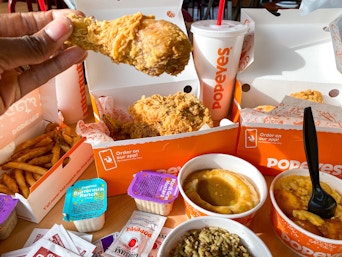A person's hand holding up a piece of chicken above a Popeyes chicken family meal displayed on a table at Popeyes.