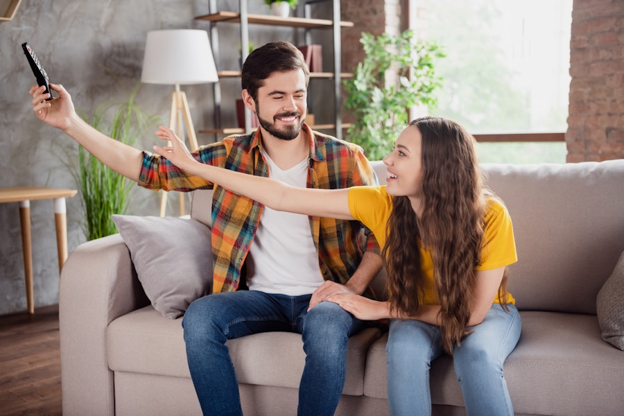 Two people sitting on a couch, one holding the TV remote away from the other who is reaching for it