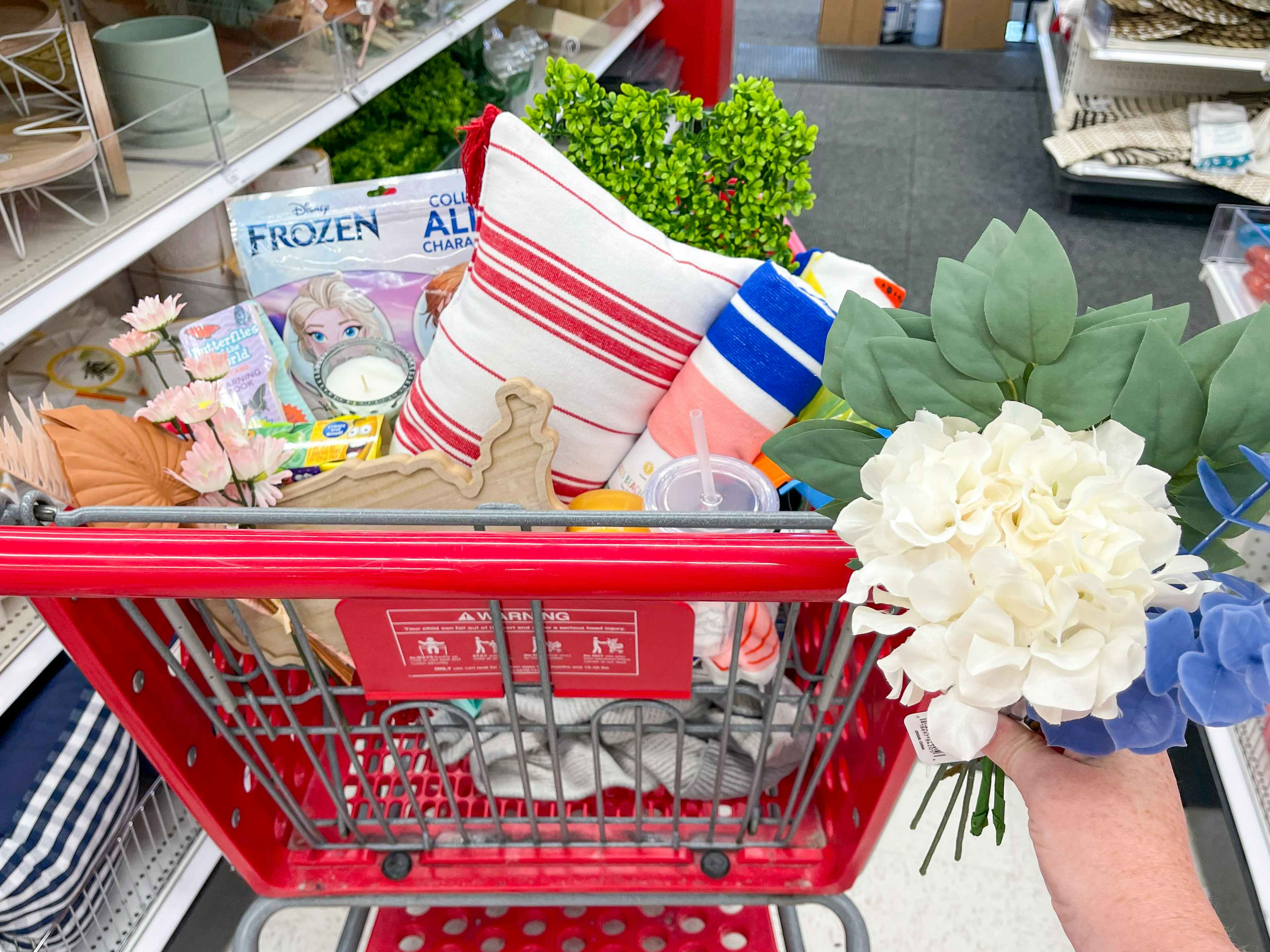 A person's hand holding up a bundle of silk flowers next to a Target shopping cart's basket filled with items from the Dollar Spot.