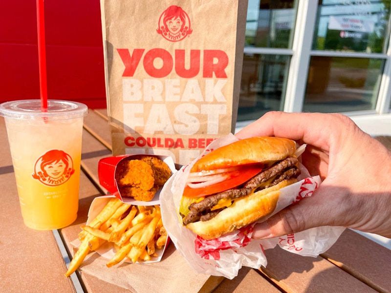 A persons hand holding a Wendy's double stack burger in front of fries, chicken nuggets, a drink, and a Wendy's bag on a table outside ...