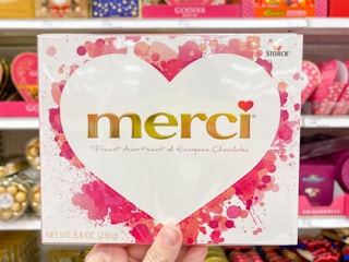 A merci assorted chocolates box held out by hand in front of a store shelf with chocolates on it.