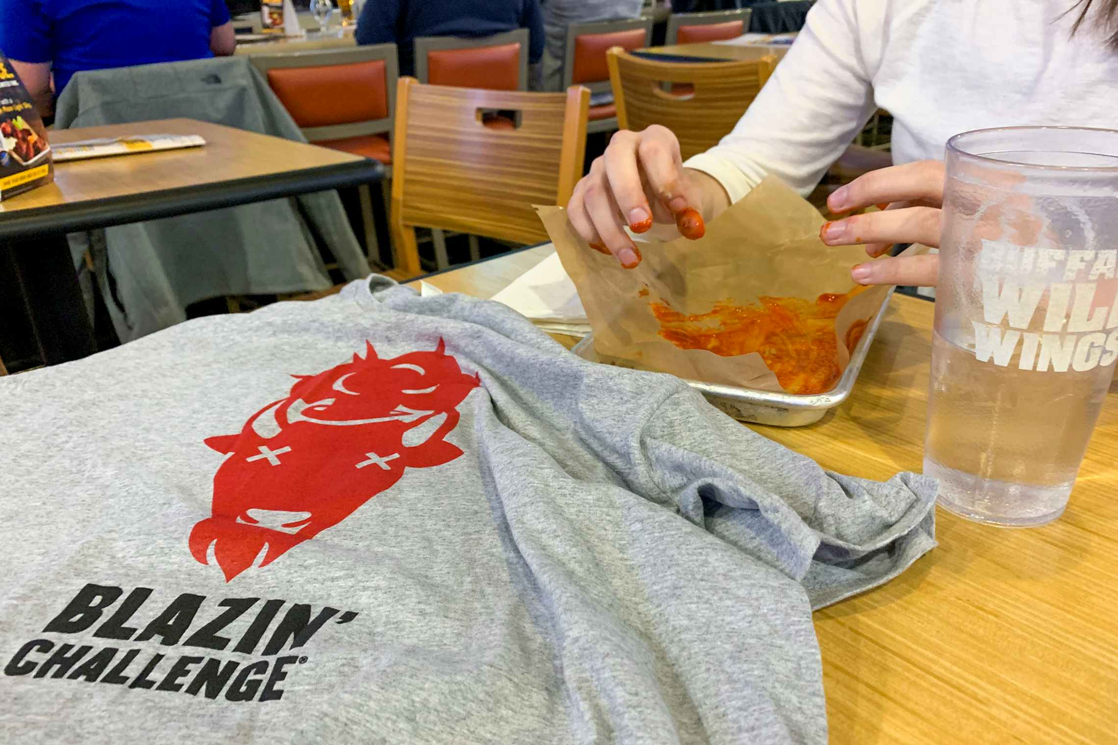 A woman sitting in a Buffalo Wild Wings restaurant with her fingers covered in buffalo sauce over an empty platter. There is a cup of wat...