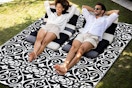 A waterproof outdoor rug in beige and black has a pattern of spirals that look like hearts in some places. Two people sit on lawn chairs