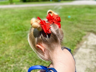 A little girl standing outside, showing off her hairstyle - two high buns with big red bows