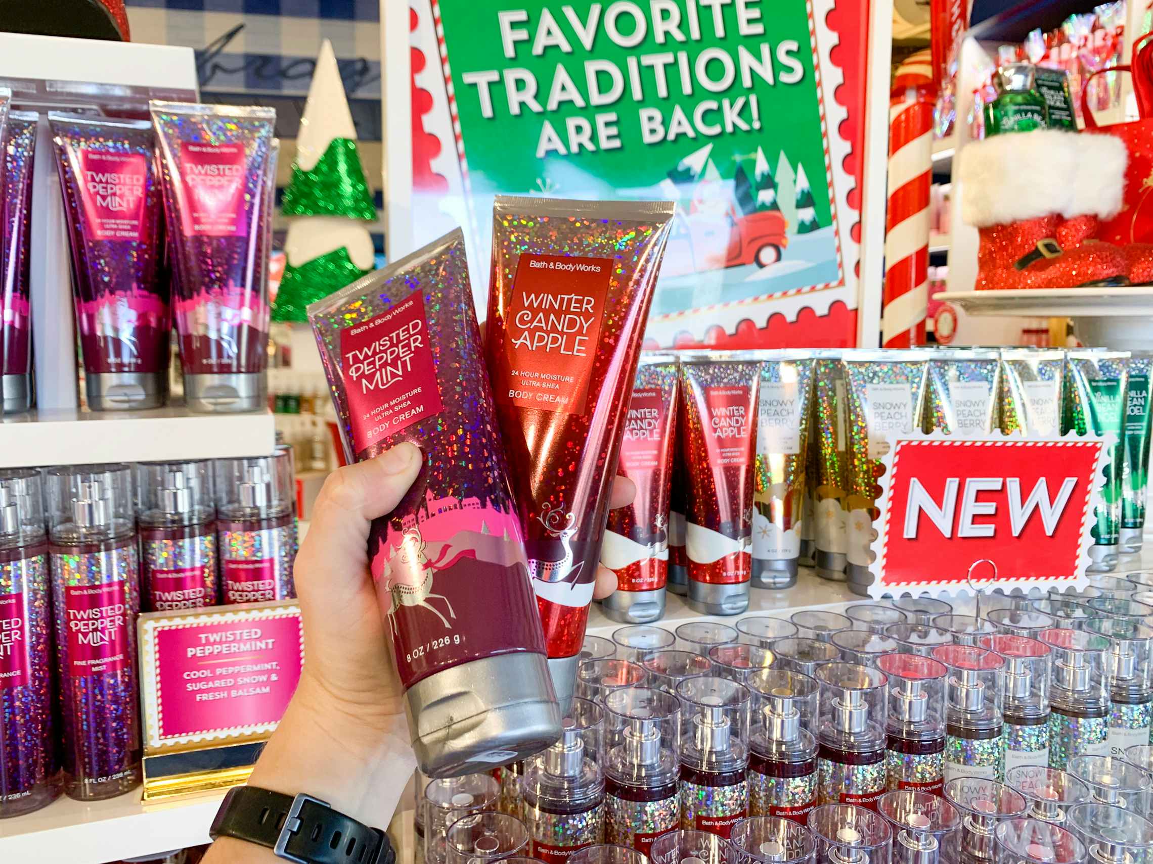  a person holding two bottles of bath and body works lotion in front of a holiday display