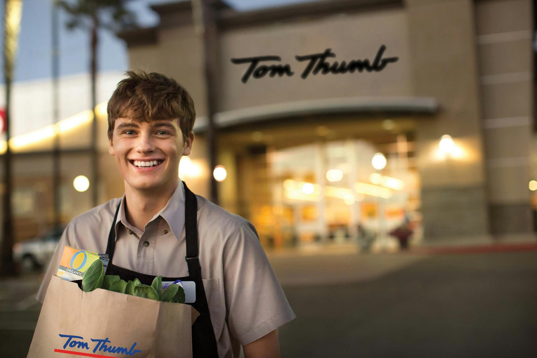 Tom Thumb store with smiling employee holding a grocery bag in front of it.