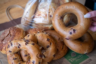 a person holding a bagel with a dozen more sitting on the table beside it.