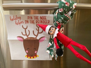 an elf on the shelf doll playing pin the nose on the reindeer holding on a kitchen towel