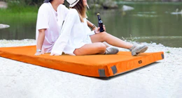 a couple sitting on a camping mattress near a lake