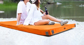 a couple sitting on a camping mattress near a lake
