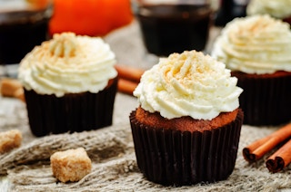 Pumpkin pie cupcakes on a table with drinks, cinnamon sticks, a pumpkin, and some crumbles.