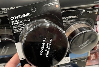 A hand holding Covergirl loose powder in a store aisle.