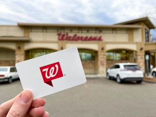 A person holding a Walgreens gift card in front of Walgreens.