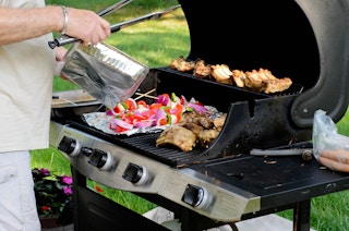 There is an open grill with ribs and vegetable skewers on tin foil cooking on it. A person is holding tongs and a foil container next ...