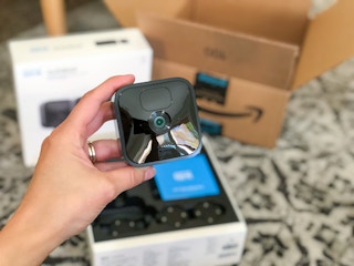 A person's hand holding a Blink outdoor wireless security camera above its box next to an Amazon delivery box.
