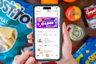 A person's hands holding a cell phone displaying the Fetch Rewards app, with groceries laid out on a table below it.