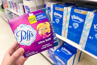 A person holding a box of Puffs facial tissues with boxes of Kleenex in the background