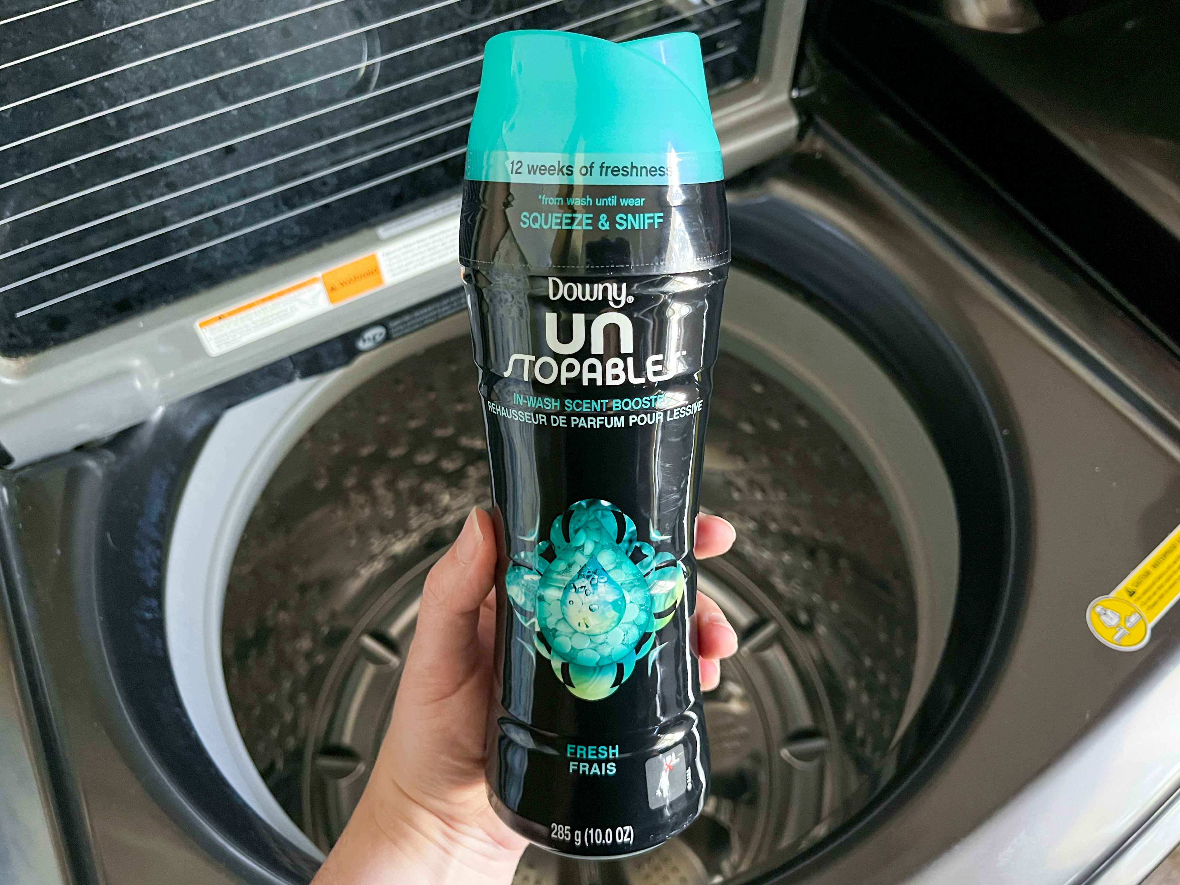 A person's hand holding a bottle of Downy Un Stopables laundry scent boost over an open washing machine.