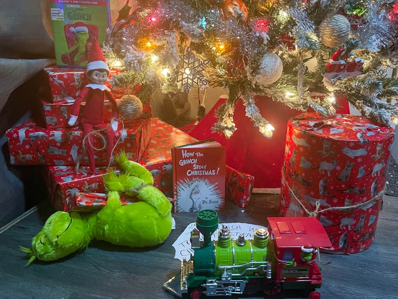 elf on the shelf sitting under a tree with wrapped gifts and a tried up grinch doll with a grinch book, movie, and train 