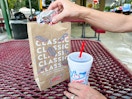 a person grabbing a hamburger out if sonic to go bag while holding a drink sitting outside sonic