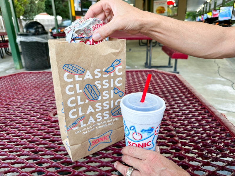 a person grabbing a hamburger out if sonic to go bag while holding a drink sitting outside sonic