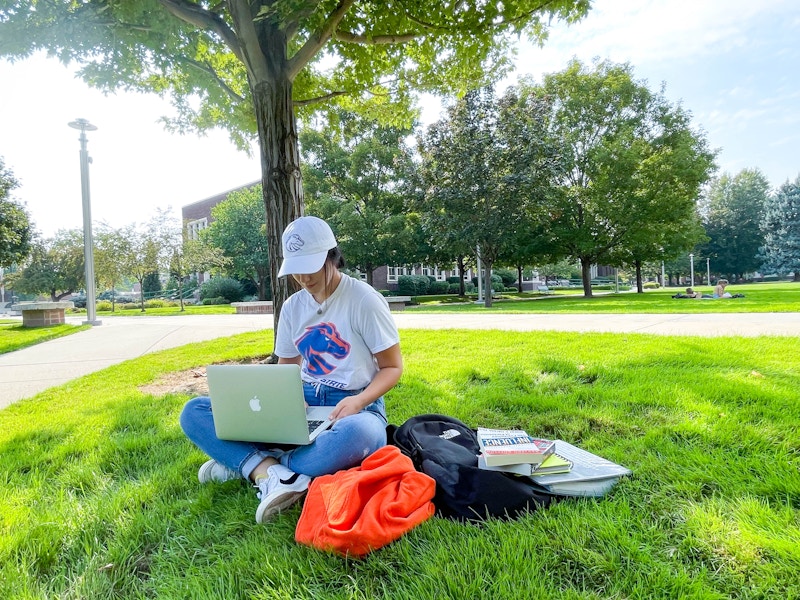 A student sitting in grass on campus with their laptop and some books.