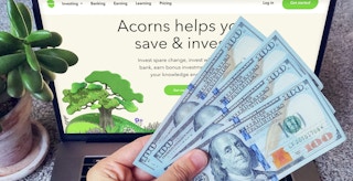 Someone holding some money in front of a laptop displaying the Acorns website