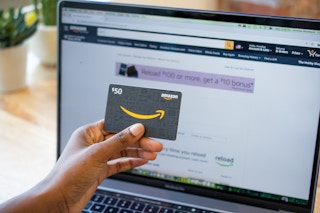 A woman holding an Amazon gift card in front of a computer screen with the Amazon gift card reload page up on it.