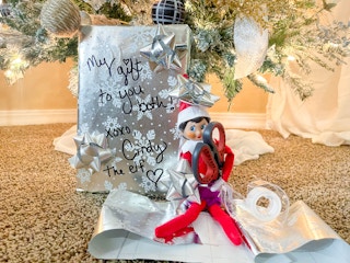 elf on the shelf doll sitting under a christmas tree next to a wrapped gift holding tape and scissors 