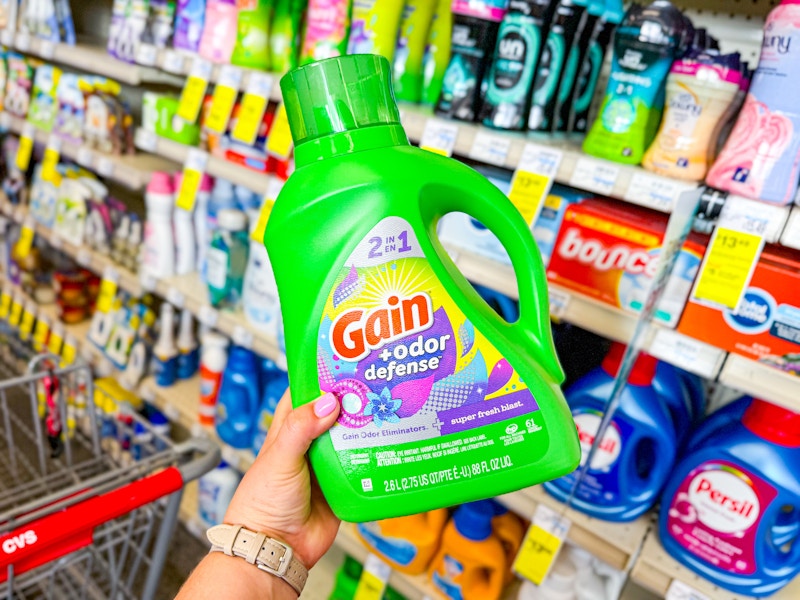 person holding a bottle of gain detergent in front of a laundry shelf