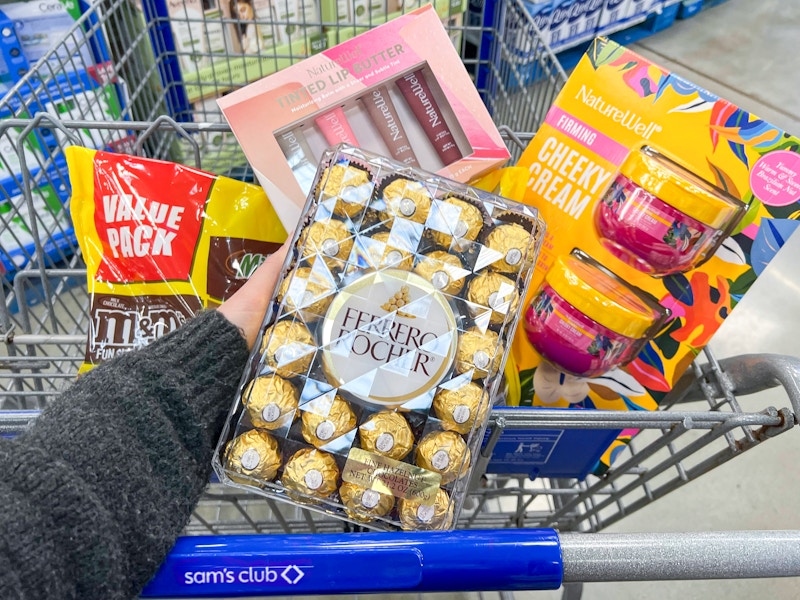 person holding a package of ferrero rocher chocolates in front of a cart with a bag of candy, lip butter, and body butter in it