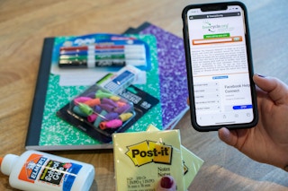 Someone holding a phone with the Freecycle website displayed on it in front of school supplies on a table.