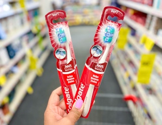 hand holding two Colgate Optic White toothbrushes in aisle