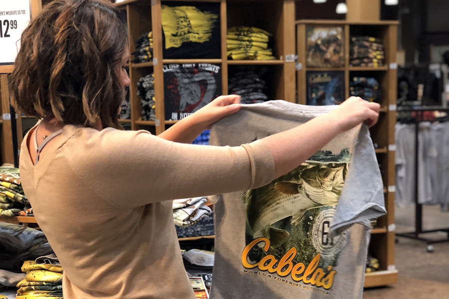 A woman holding up and looking at a Cabela's t-shirt in the clothing section of Cabela's.