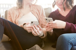 two women from the neck down sit on a couch and show each other their phones