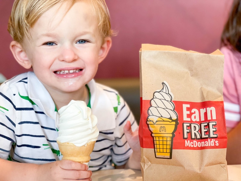 A child sitting in a booth at McDonald's, holding a soft serve ice cream cone next to a takeout bag that says, "Earn Free McDonald's