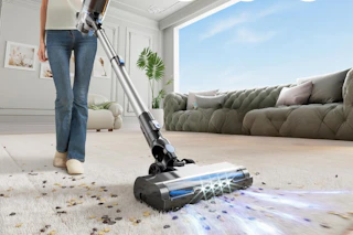 A person in jeans pushes the cordless vacuum cleaner with headlights to illuminate mess, across a carpeted floor full of bits of something.