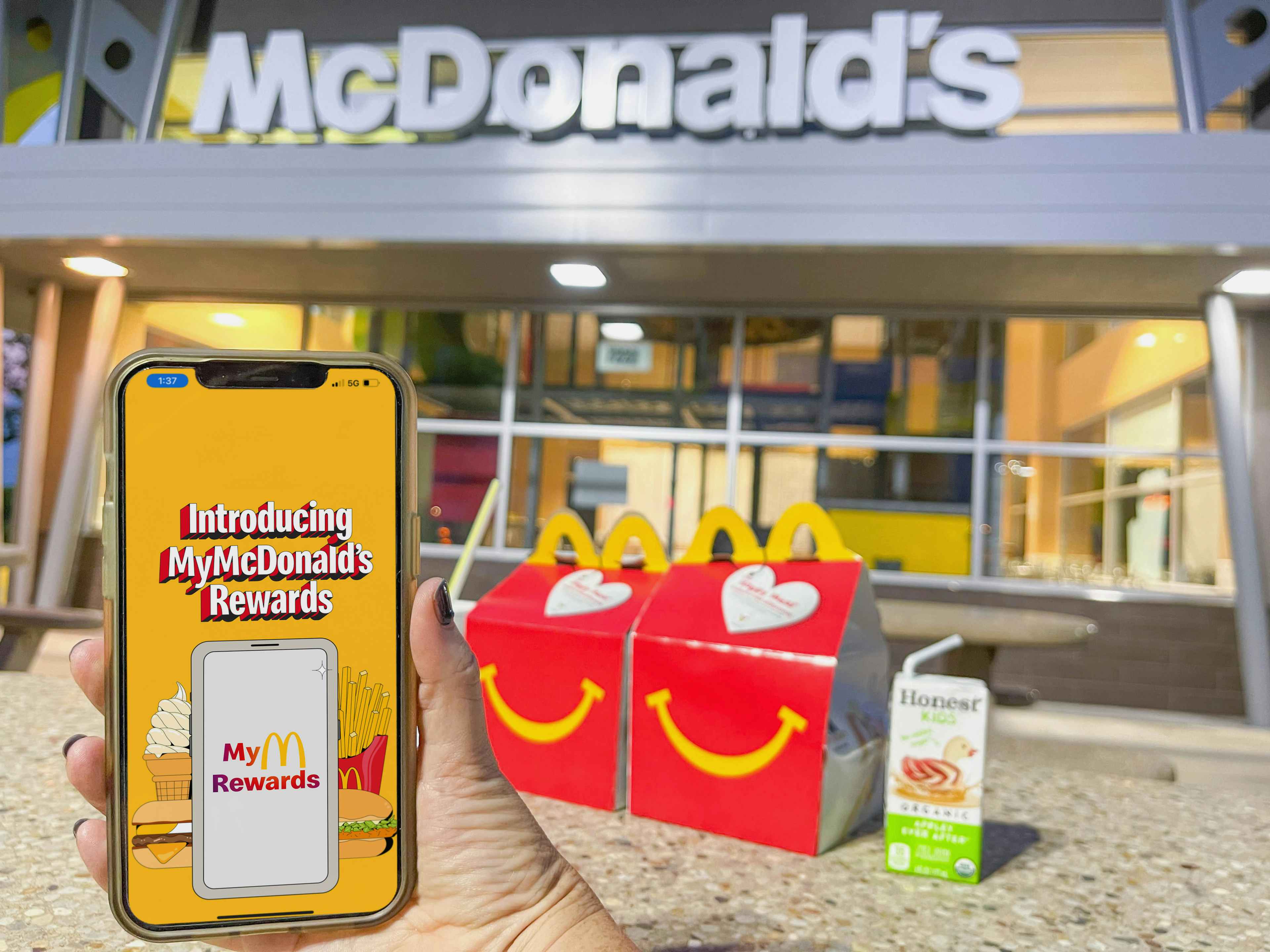 A person's hand holding up a cellphone displaying the McDonald's Rewards app in front of two Happy Meals and a juice box sitting on a tab...