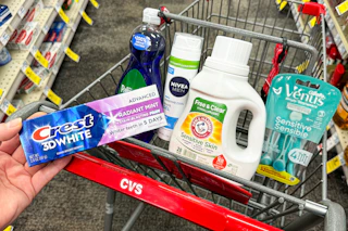 person holding crest toothpaste over a cart with palmolive dish soap, nivea shave gel, arm & hammer detergent, and venus razors in it