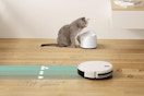 A robot vacuum and mop vacuums and mops a hardwood floor. Aqua lines and white droplets represent this. Nearby a messy cat eats.