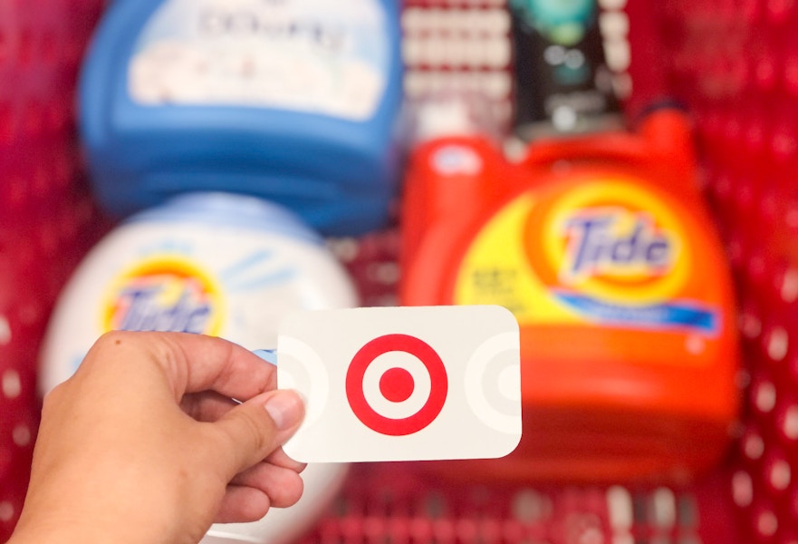 A person holding gift card over the top of a shopping cart that has bottles of laundry detergent