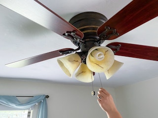 A person's hand reaching up to pull the chain switch on a ceiling fan.