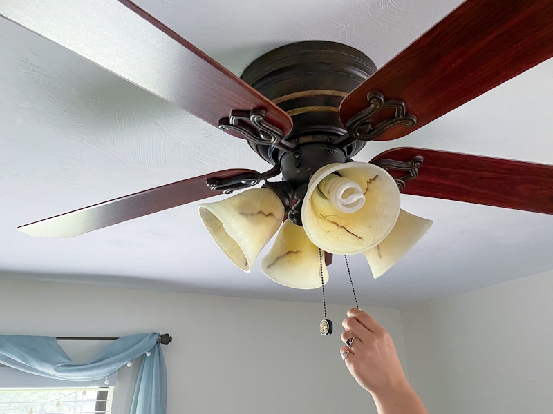 A person's hand reaching up to pull the chain switch on a ceiling fan.