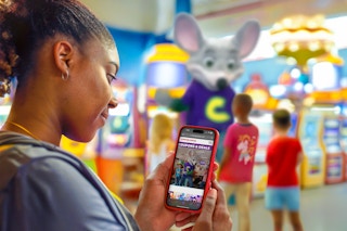 a woman looking at coupons for chuck e cheese on her phone, kids in the background interacting with the chuck e cheese mascot in the arcade