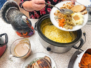 A plate of Thanksgiving dinner on a plate in front of a crock pot with creamed corn being scooped out on the plate.