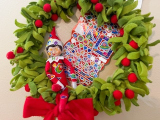 an elf on the shelf doll sitting in a felt wreath covered in stickers with a sticker book behind elf 