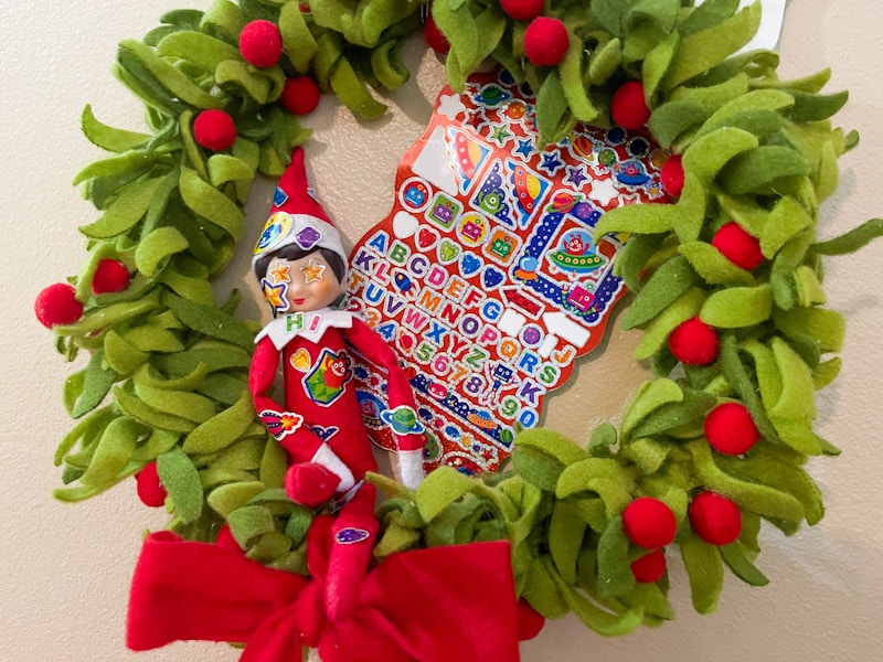 an elf on the shelf doll sitting in a felt wreath covered in stickers with a sticker book behind elf 