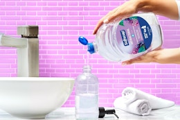 In a pink bathroom, two hands pour a Softsoap White Tea and Berry refill into a sink dispenser.